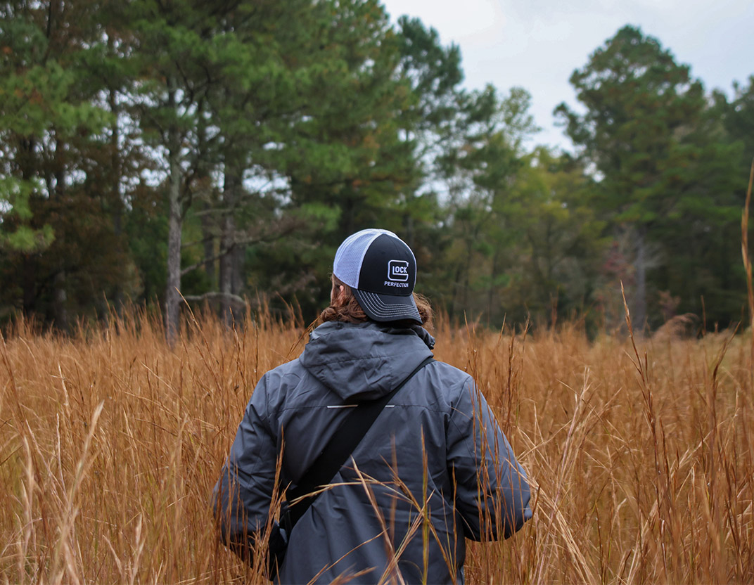 Man walking in grass with back to camera, wearing a Glock hat